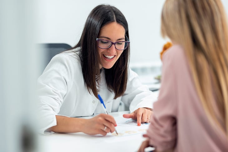 doctora joven sonriendo, escribiendo en una hoja, en consulta con mujer-1268569670 (2)